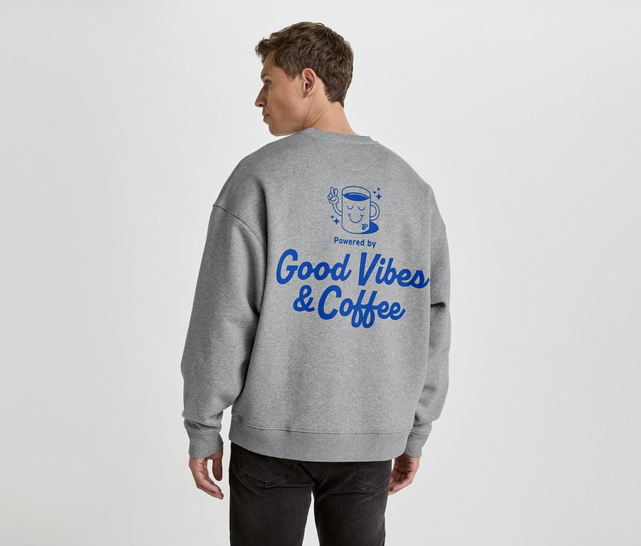 Hátulról egy férfi látható szürke pulóverben, kék 'Good Vibes & Coffee' felirattal.