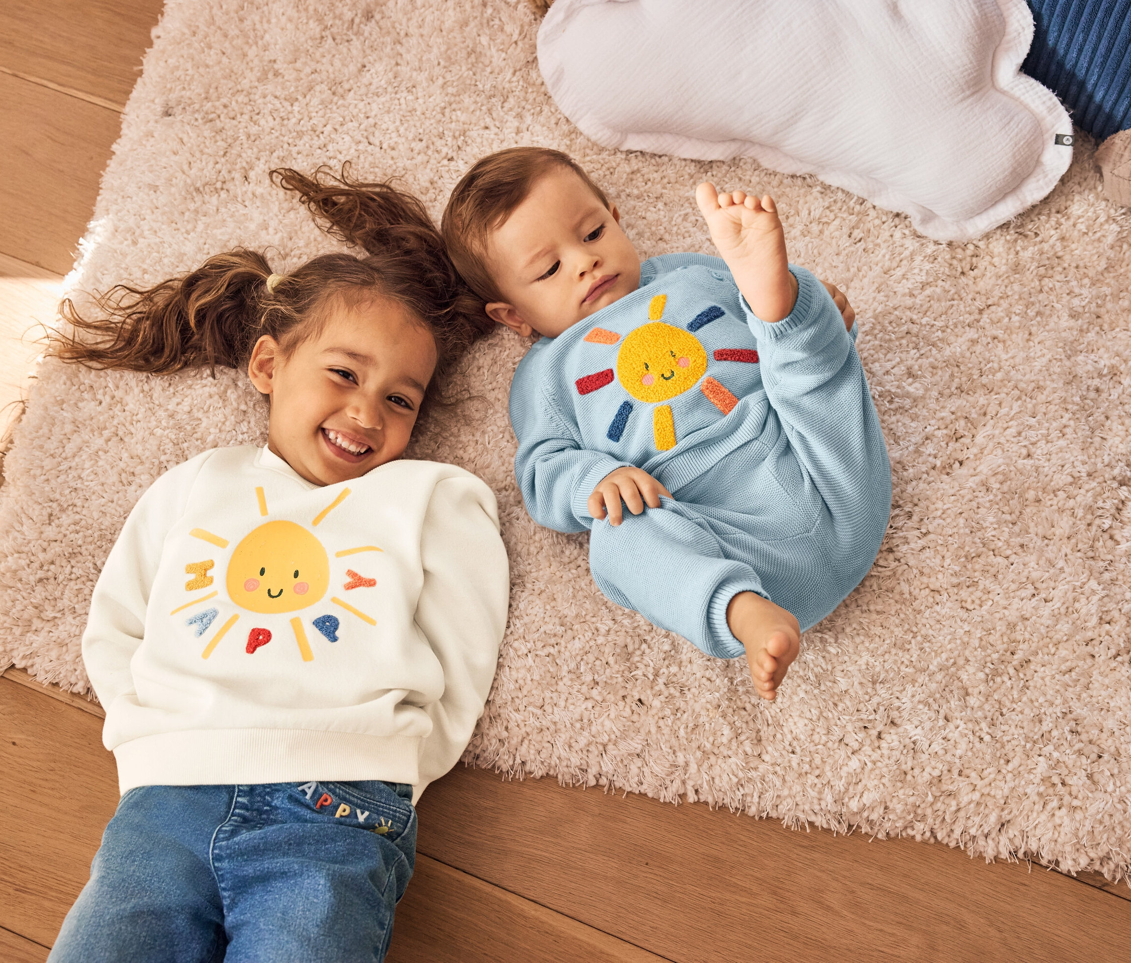 Egy copfos kislány és egy baba fekszik egy szőnyegen. A lány egy sárga napos krémszínű pulóvert visel. A baba kék kötött ruhát visel, napocskával.