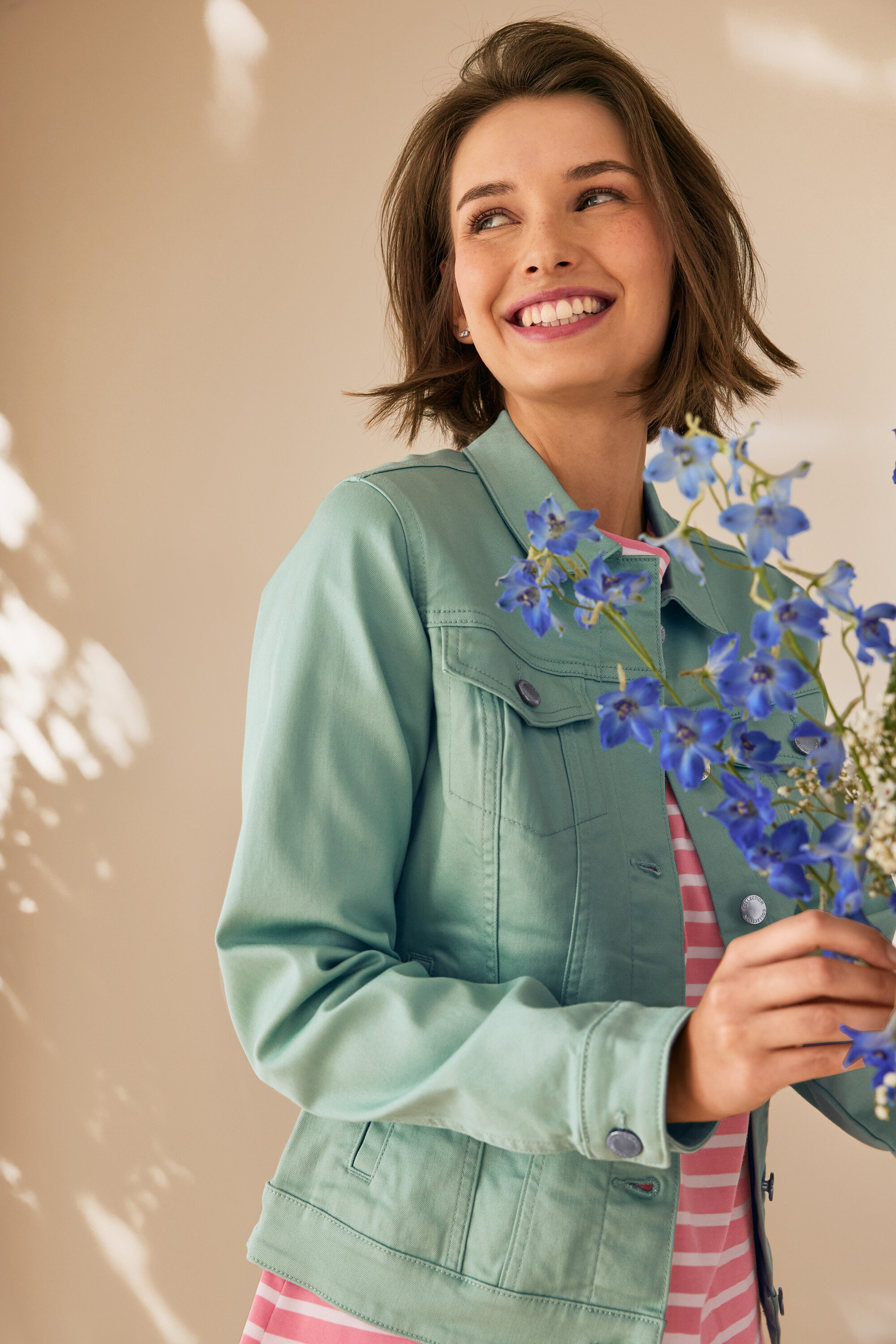 Mosolygó nő kék virágokat tart, mentazöld farmerdzsekit visel.