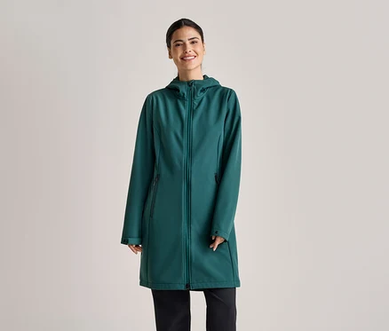 A lófarokba kötött hajú nő smaragdzöld, kapucnis, hosszú softshell kabátot és fekete nadrágot visel világosszürke háttér előtt.