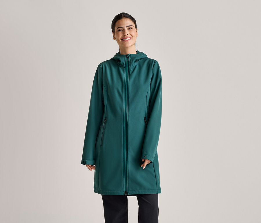 A lófarokba kötött hajú nő smaragdzöld, kapucnis, hosszú softshell kabátot és fekete nadrágot visel világosszürke háttér előtt.