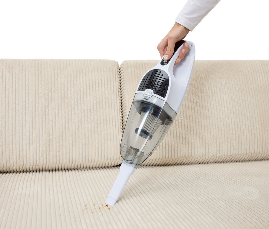 Egy kéz morzsákat porszívózik fel egy bézs kanapéról egy 2 az 1-ben akkumulátoros porszívóval.