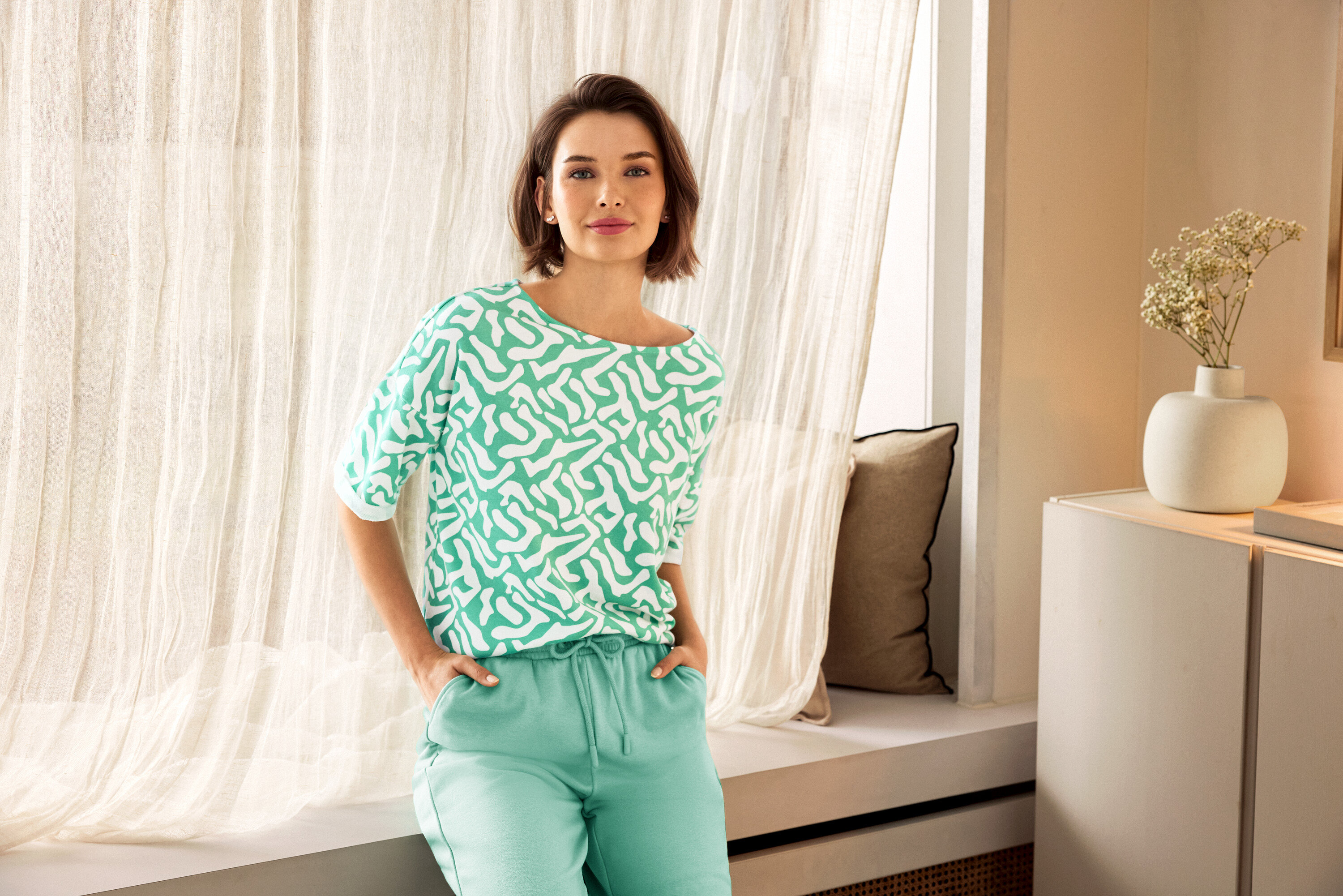 Nő zöld nadrágban és mintás felsőben pózol egy ablak mellett. A háttérben egy kerámia váza látható.