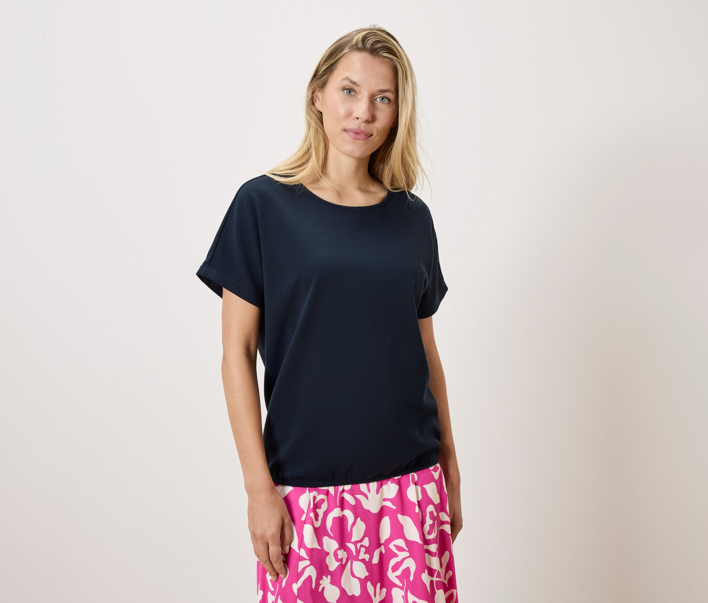Egy szőke nő sötétkék pólót és rózsaszín-fehér mintás szoknyát visel fehér háttér előtt.