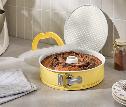 Márványos sütemény sárga tortaformában fedővel és emelőfogantyúval, konyhai robotgép és fűszertartó üveg mellett
