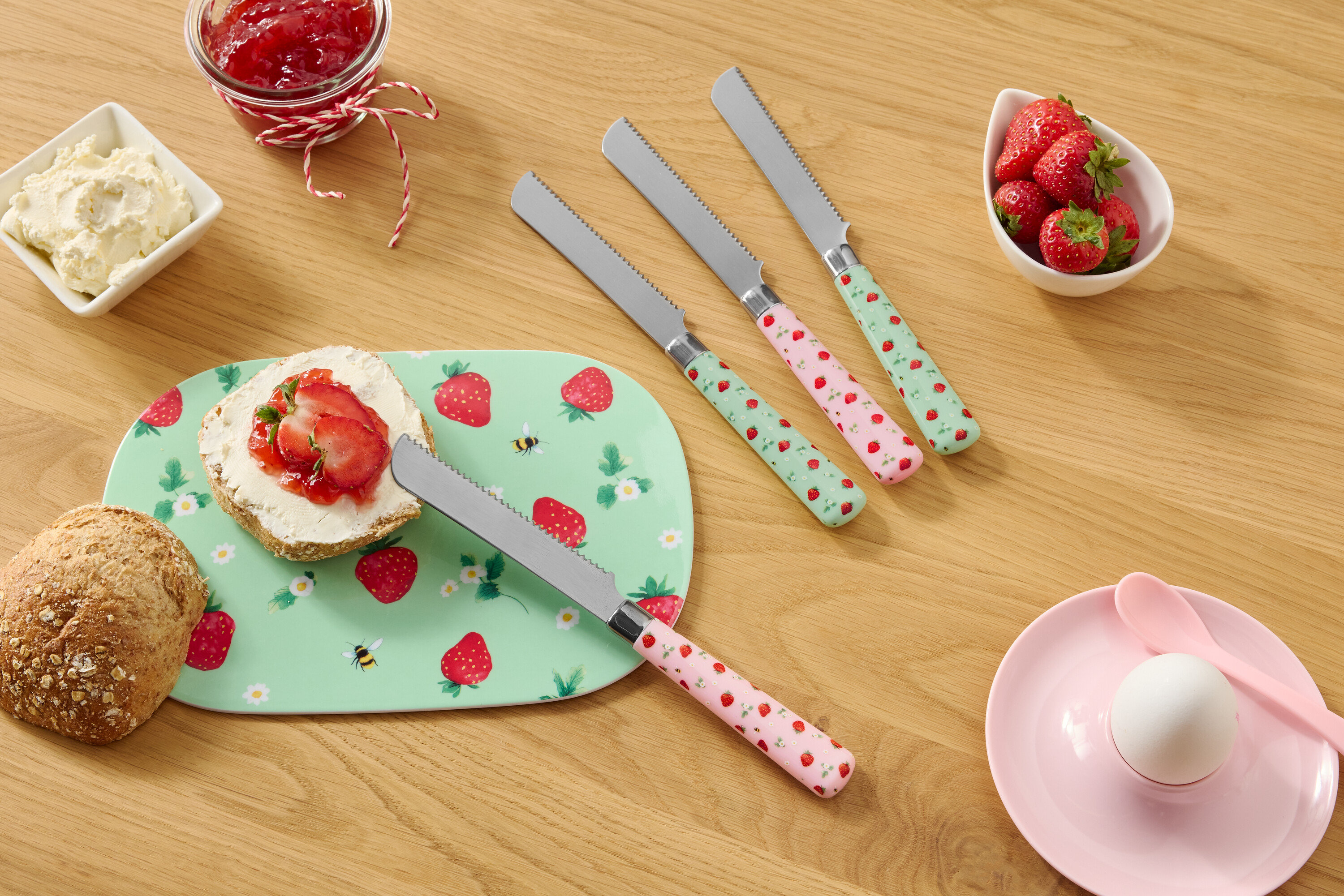 Egy fa asztalon egy zsemle, egy eper mintás tányér, krémsajt, lekvár, eper és négy eper mintás kés található.