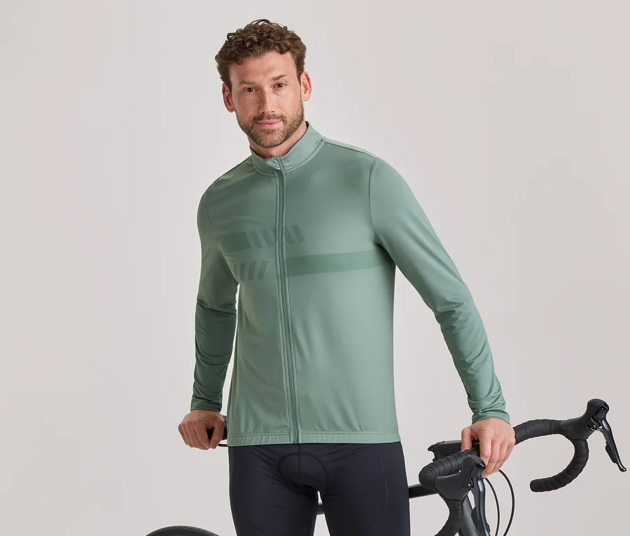 Egy rövid göndör barna hajú, enyhe szakállú férfi zsályazöld hosszú ujjú kerékpáros dzsekit visel finom mintával és fekete kerékpáros nadrágot, állva tartja egy fekete országúti kerékpár kormányát, és a kamerába néz.