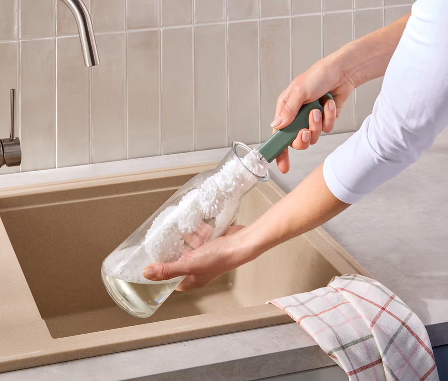 Egy személy üvegpalackot tisztít zöld palackmosó kefével egy bézs mosogató felett. Egy kockás konyharuha lóg a mosogató szélén.