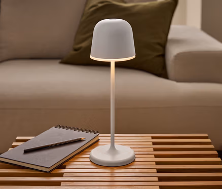 Egy világító fehér Eglo »Mannera« LED-es, akkumulátoros asztali lámpa áll egy léces faasztalon, jegyzetfüzet és ceruza mellett, egy kanapé előtt.