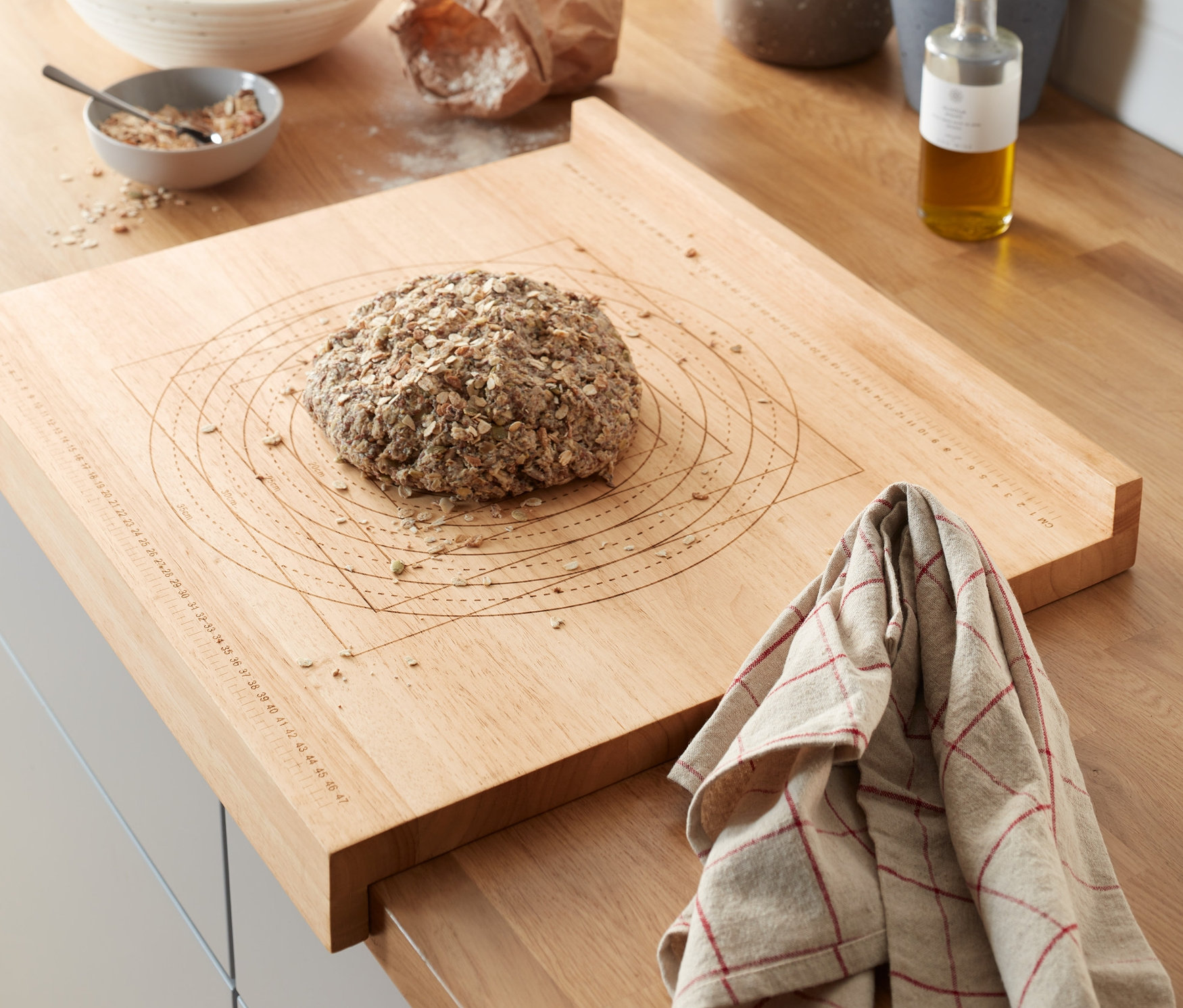 A konyhapulton egy fából készült nyújtódeszka méretskálával, egy kenyérrel. A háttérben cserepes fűszernövények és egyéb konyhai eszközök láthatók.