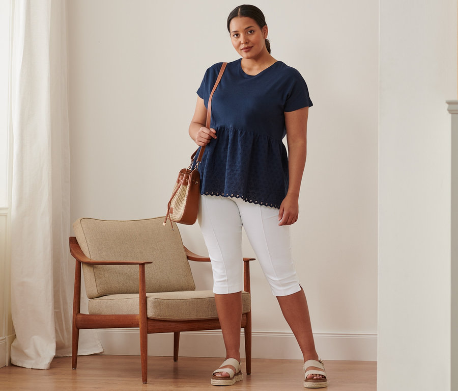 Egy nő áll sötétkék, hímzett női felsőben, 3/4-es sztreccsnadrágban, bőr szandálban és oldaltáskában.