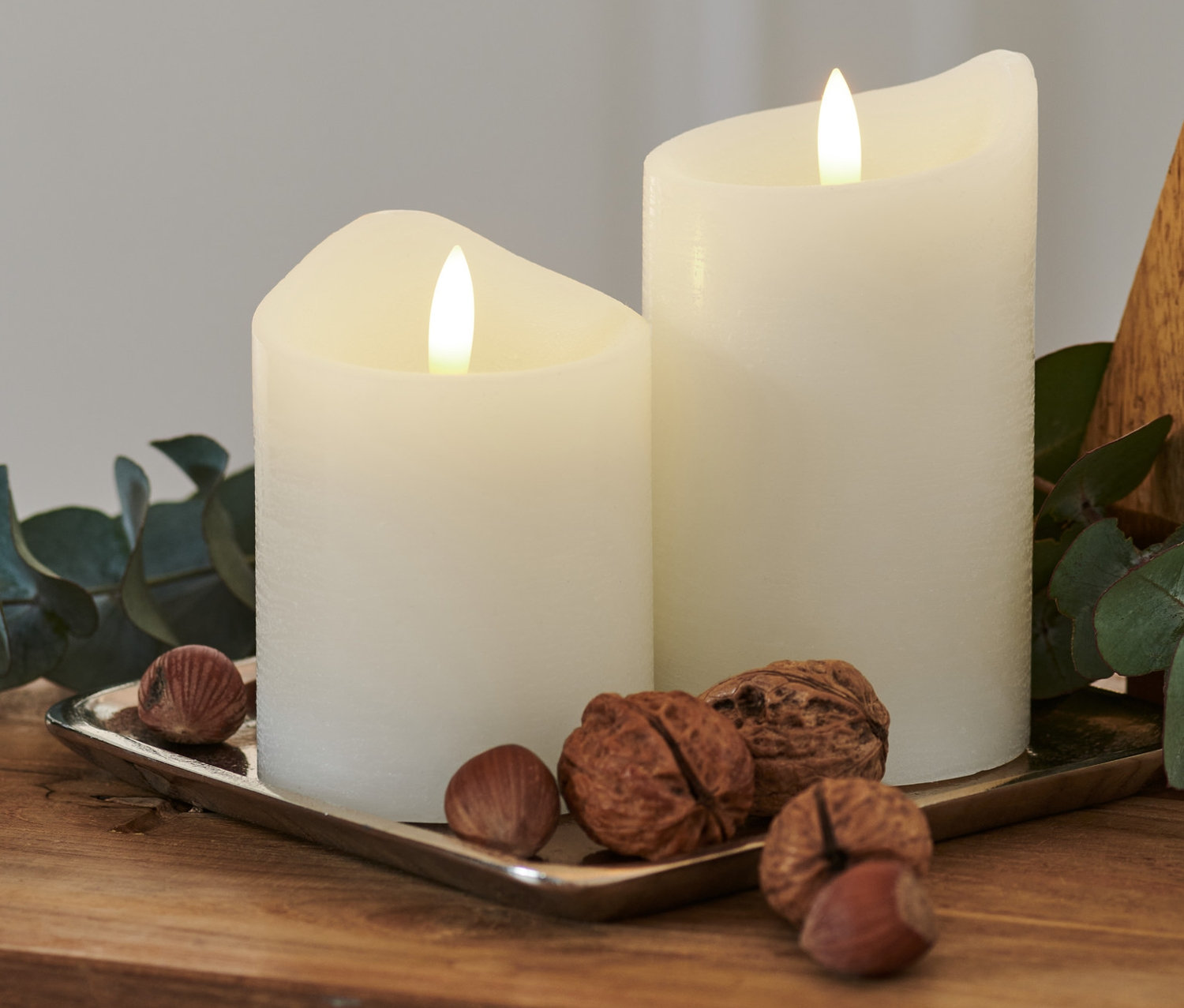 Egy fa asztalon, egy ezüst tálcán két fehér LED-es valódi viaszgyertya, diófélék és egy eukaliptusz ág látható.