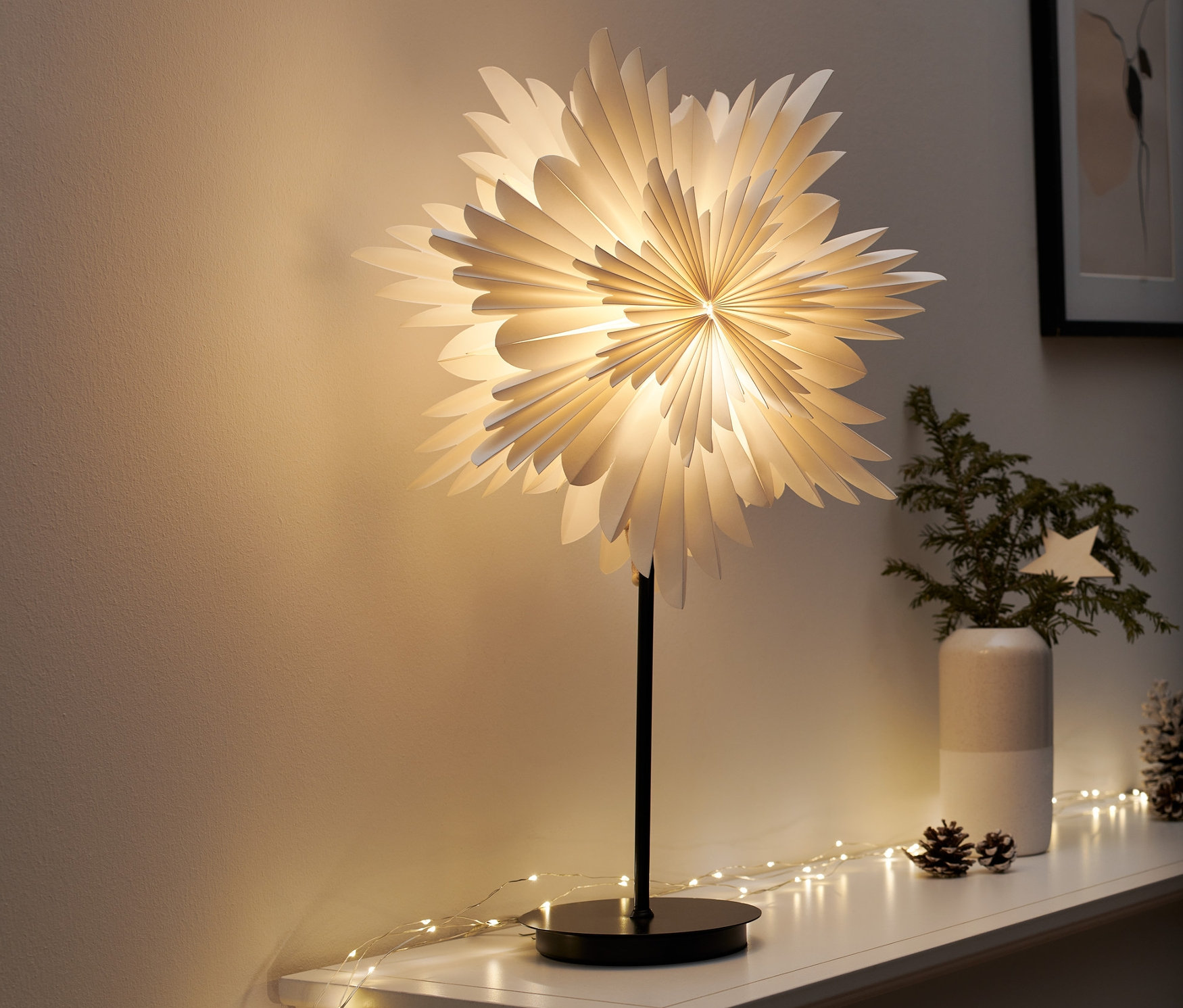 Egy LED-es csillag alakú díszlámpa áll egy fehér polcon. Körülötte fényfüzérek és karácsonyi dekorációk találhatók.