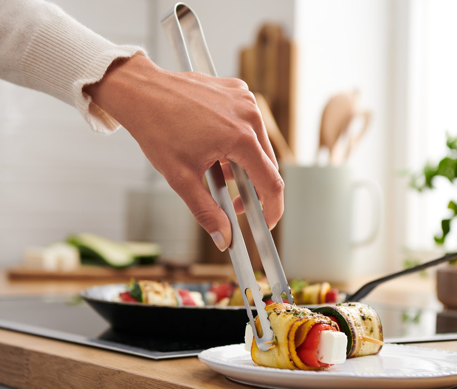 Egy kéz egy nemesacél konyhai fogóval megfog egy zöldséggel töltött cukkini tekercset egy tányéron.