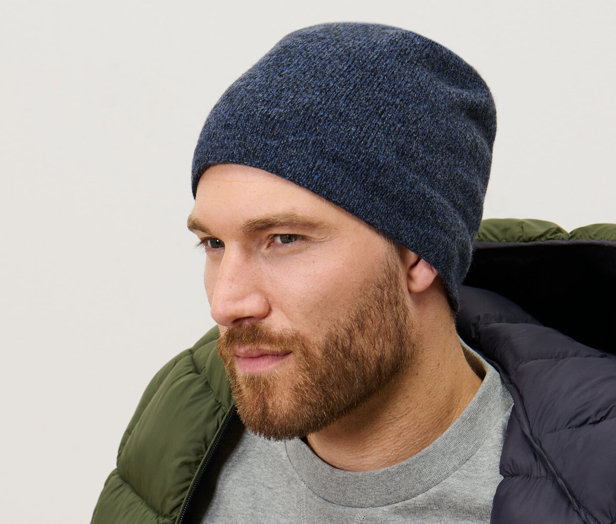 Férfi kötött sapkát, melírozott sötétkéket, szürke pulóvert és zöld színű pehelykabátot visel.