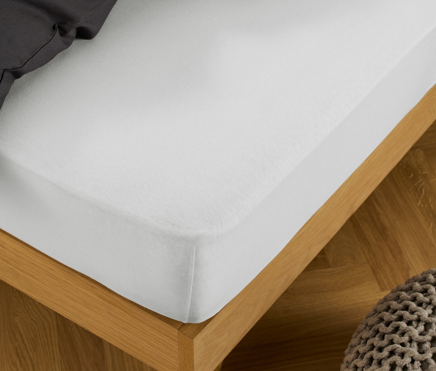 Egy fehér gumis jersey lepedő van egy fa keretes ágyon, mellette egy kötött puff.