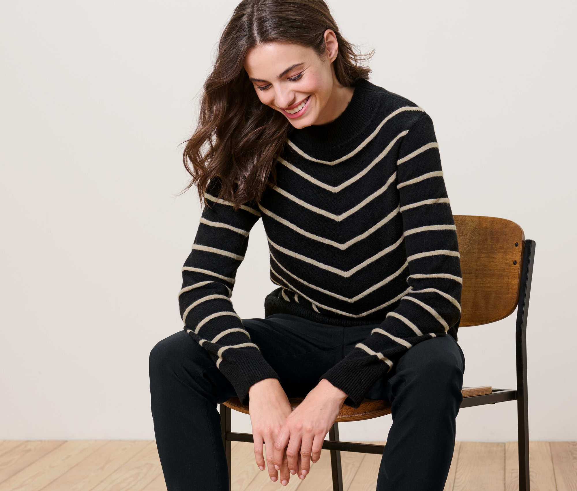 Nő ül egy széken, és fekete, csíkos kötött pulóvert visel állógallérral.