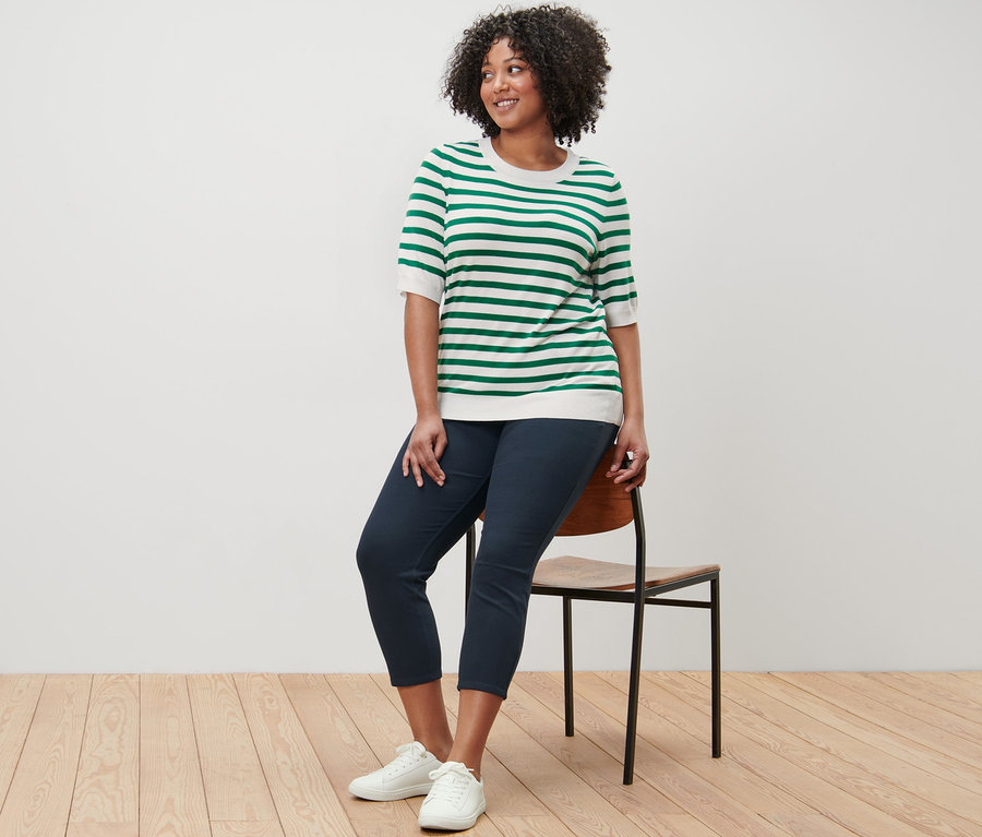 Nő ül egy széken, finom kötésű csíkos pulóvert és 7/8-os jeggingst visel.