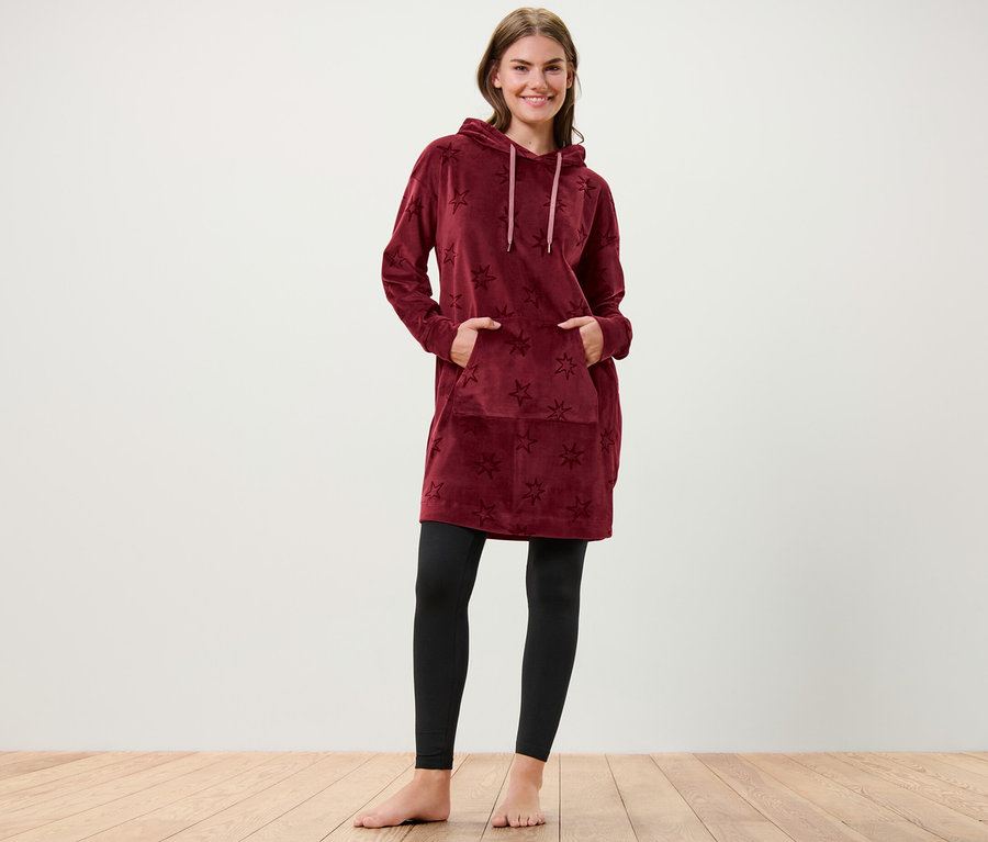 Nő áll csillagos plüss kapucnis lezser ruhában, kezét a zsebébe teszi.