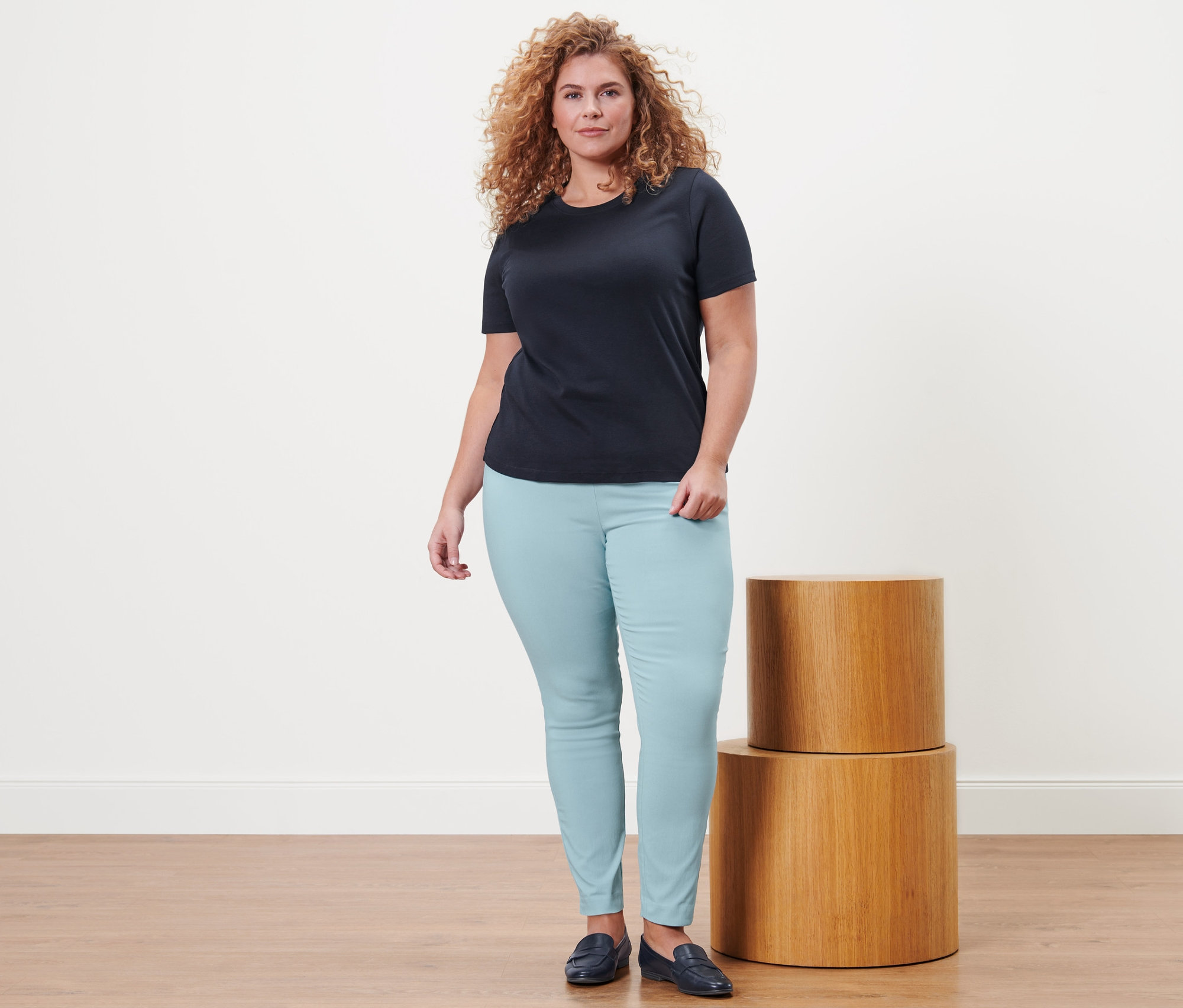 Nő áll sötétkék Basic pólóban, sztreccsnadrágban és bőr mokaszinban.