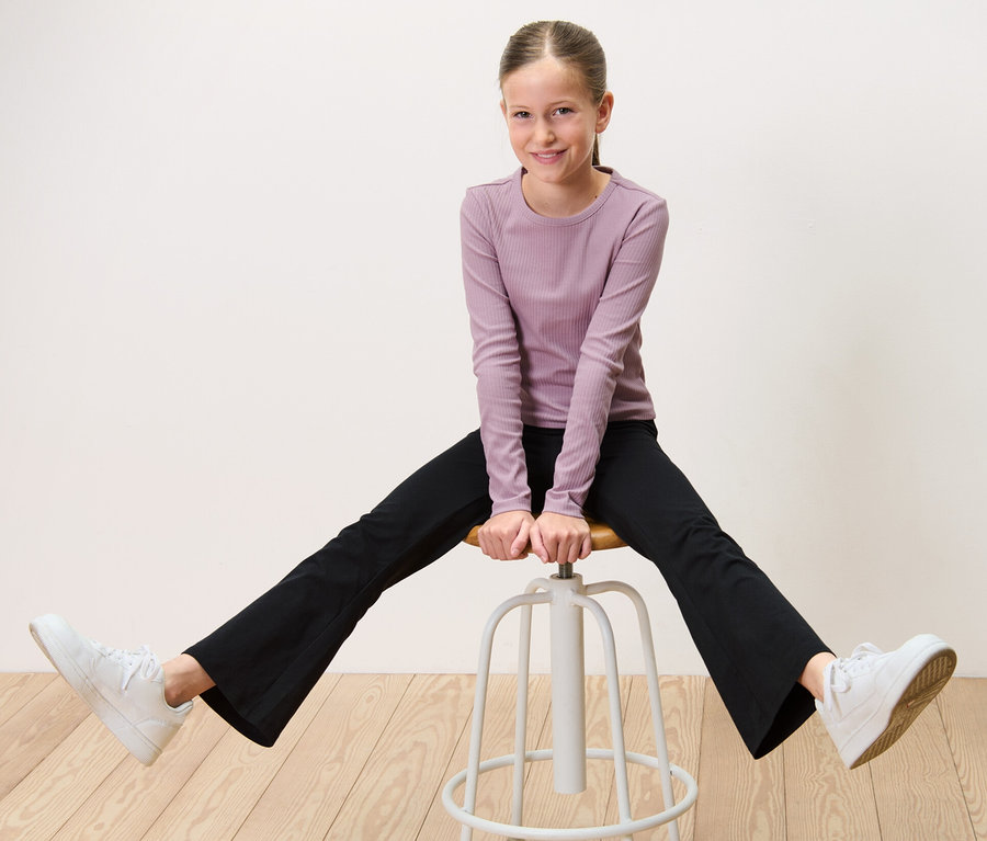 Lány terpeszben ül egy széken és fekete 2 lány trapéz leggingst visel.