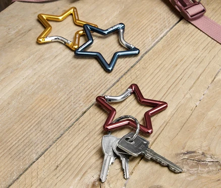 Három csillag alakú karabiner fekszik egy fa asztalon: egy arany, egy kék és egy piros, a piros karabineren kulcsok vannak. A háttérben egy hátizsák egy része látható.