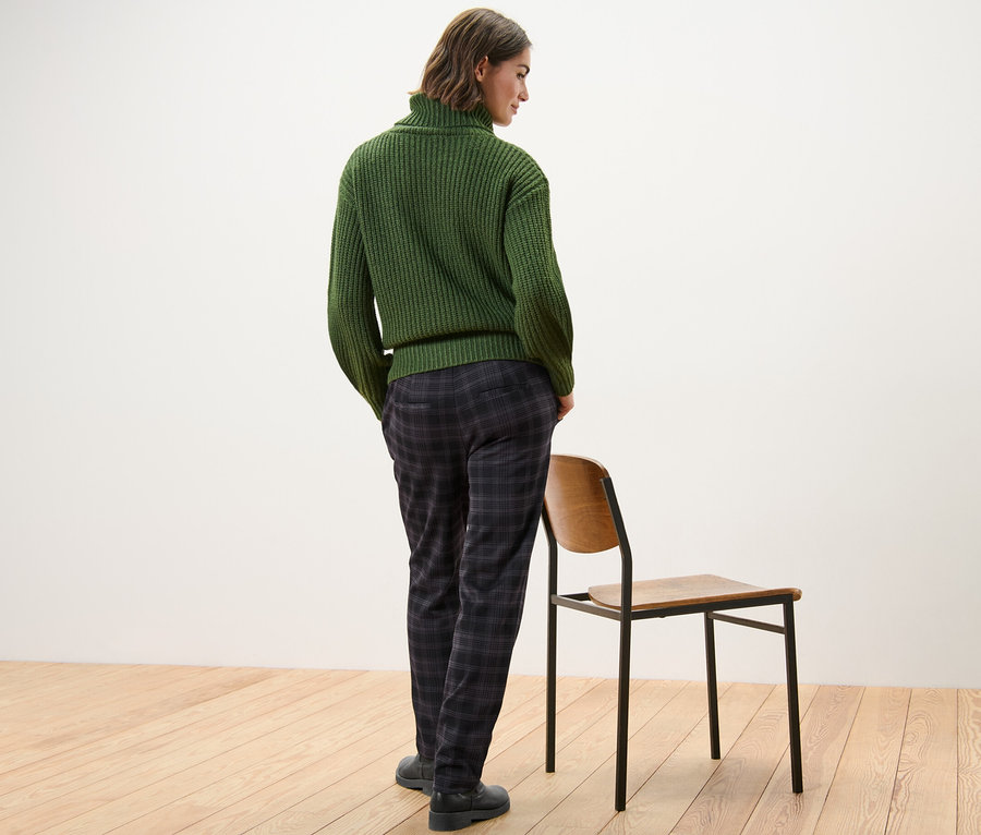 Egy nő háttal áll a kamerának, zöld pulóvert és kockás női lezser nadrágot visel.