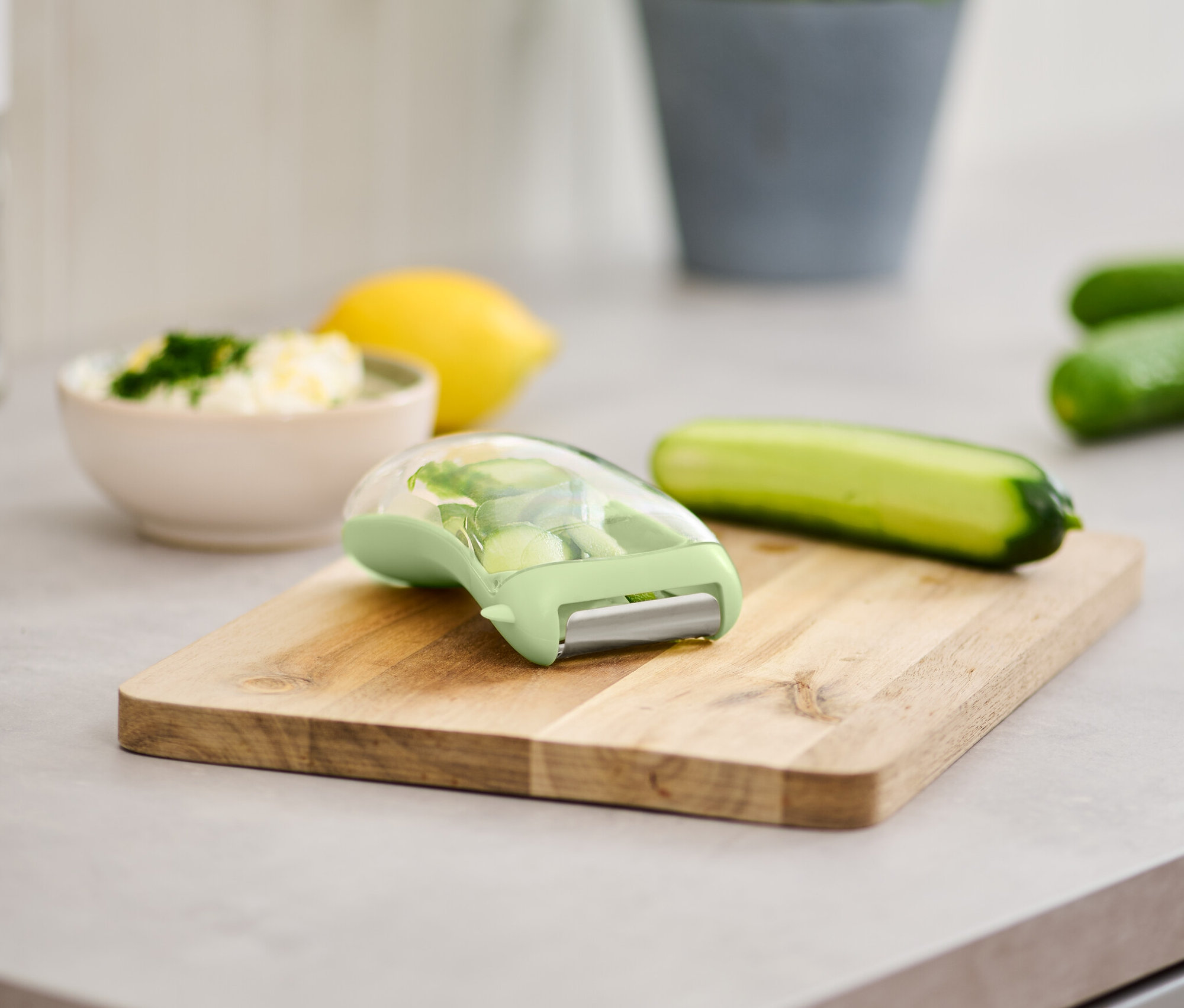 Univerzális hámozó gyűjtőtartállyal egy fa deszkán, uborka és egy tál étel mellett.