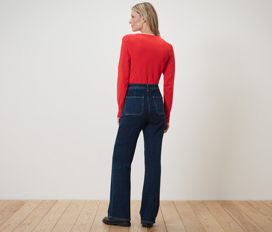 Egy nő háttal áll a kamerának, bőszárú farmernadrágot – Fit »Cara« és egy piros felsőt visel.