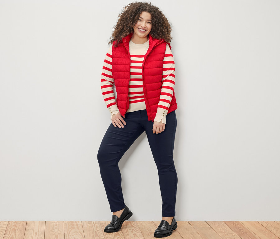 Egy nő áll és mosolyog a kamerába, piros csíkos, finomkötésű pulóvert, piros, kapucnis steppelt mellényt és sötétkék bengalin nadrágot visel.