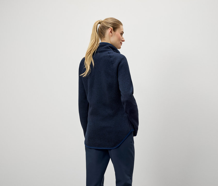 Hátulról látható egy szőke hajú, lófarokba kötött nő, aki sötétkék pulóvert és nadrágot visel.