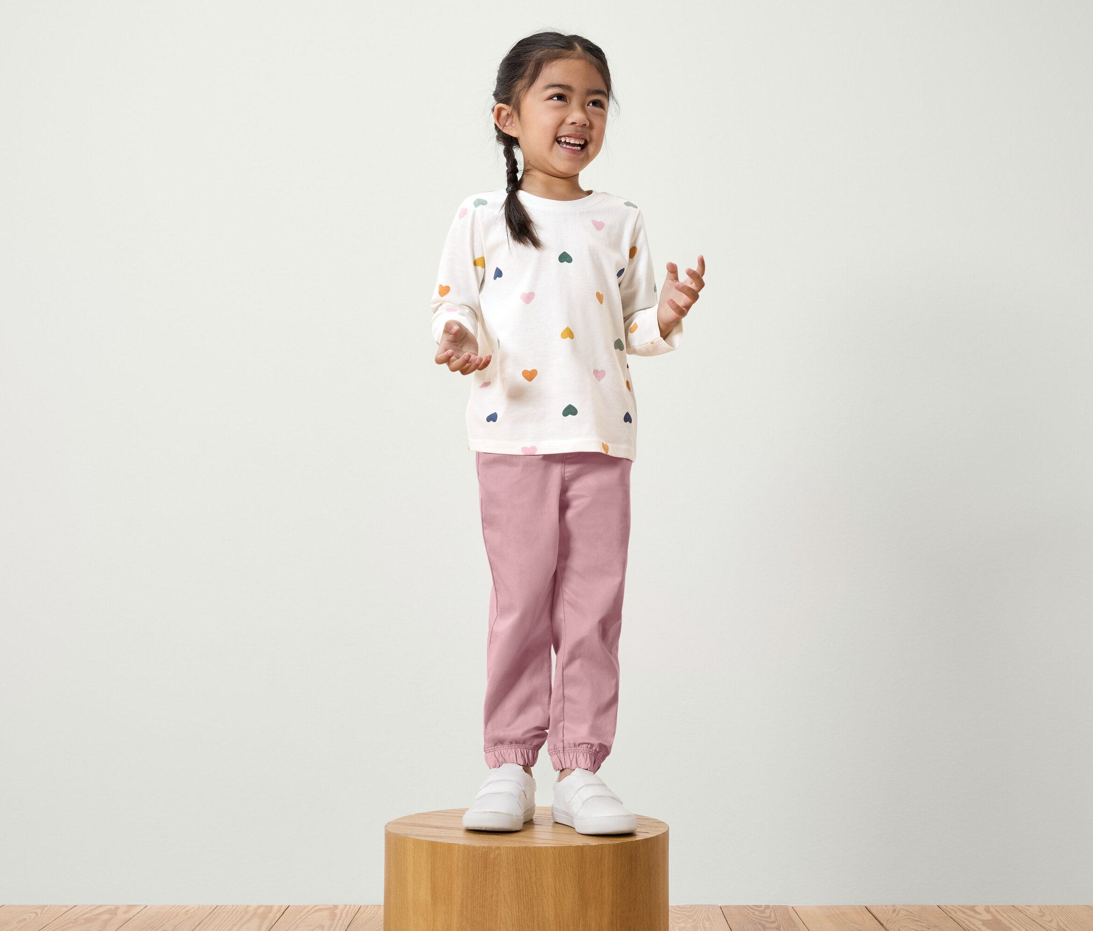 Egy copfos kislány áll egy fa talapzaton. Fehér, hosszú ujjú, színes szívecskés pólót és rózsaszín nadrágot visel.