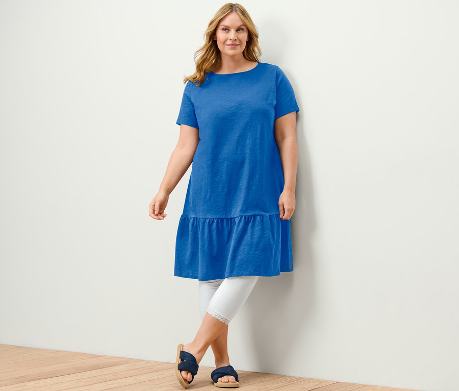 Nő pózol kobaltkék női jerseyruhában, fehér leggingsben és kék szandálban.