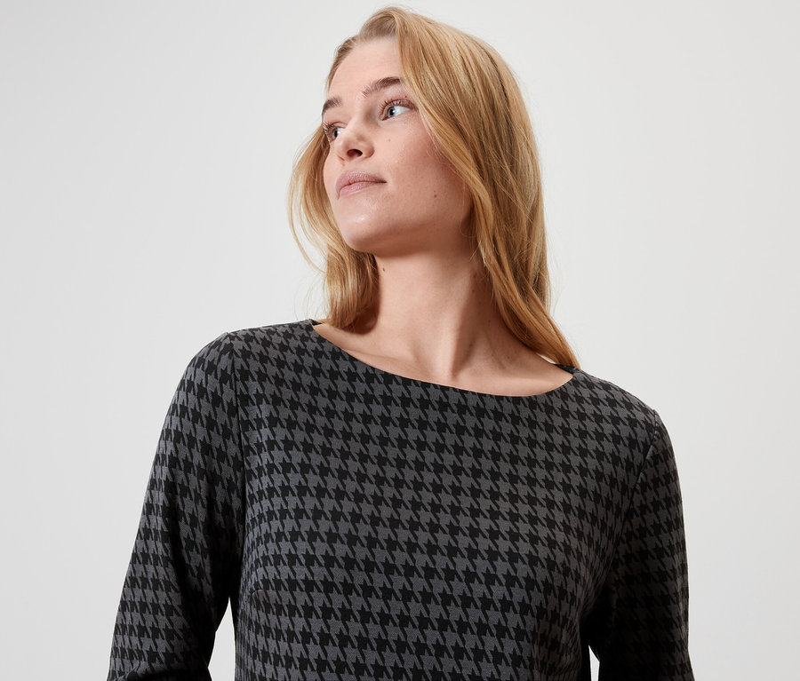 Egy szőke, hosszú hajú nő fekete-szürke, kígyóbőr mintás ruhát visel.