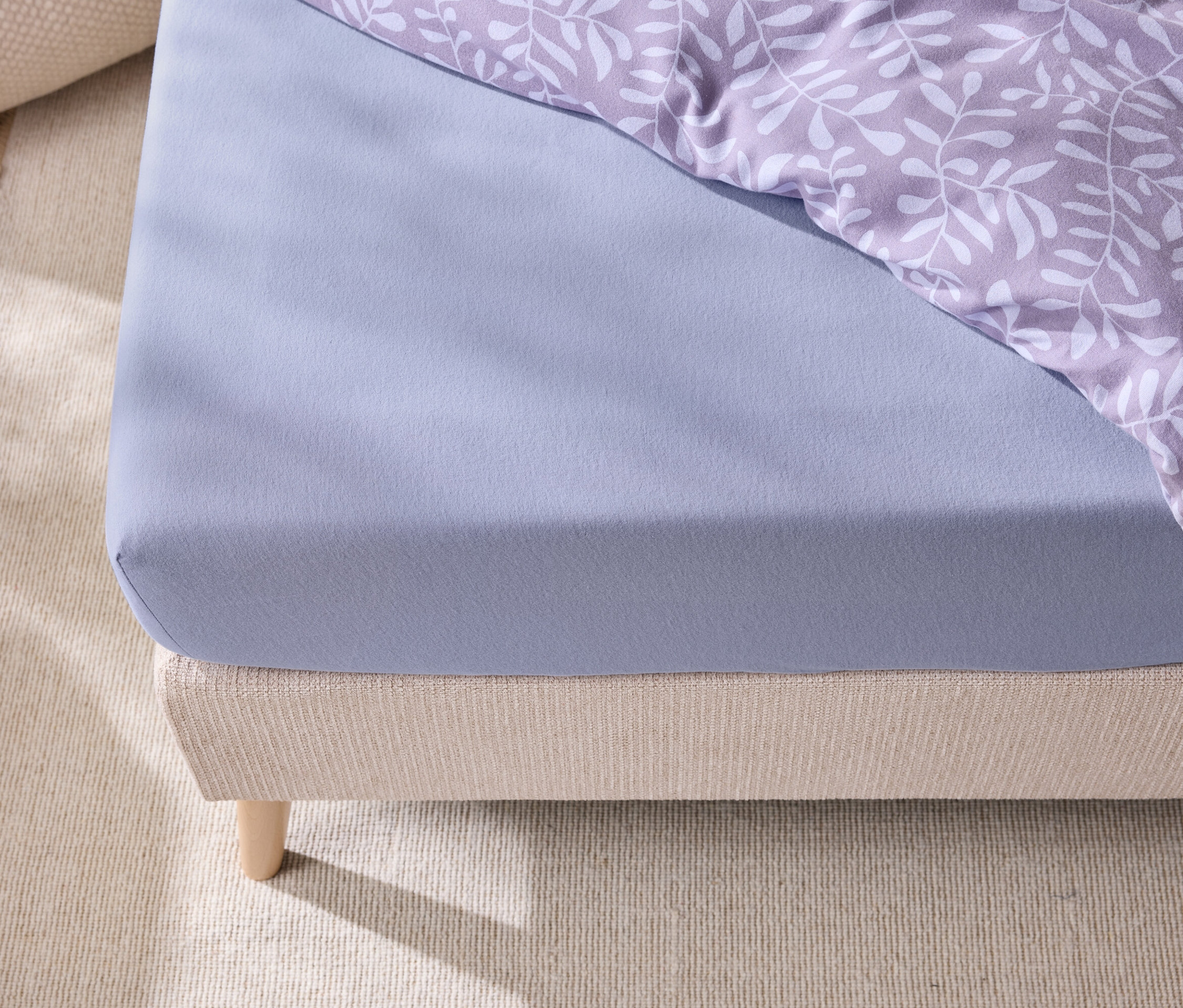 Egy ágy részlete kék gumis lepedővel és levélmintás ágyneművel.