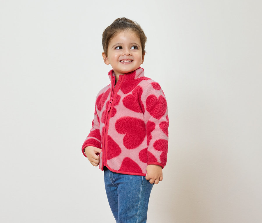 Egy lány rózsaszín, szívecskés mintázatú polár pulóvert és kék farmert visel, mosolyog és oldalra néz.