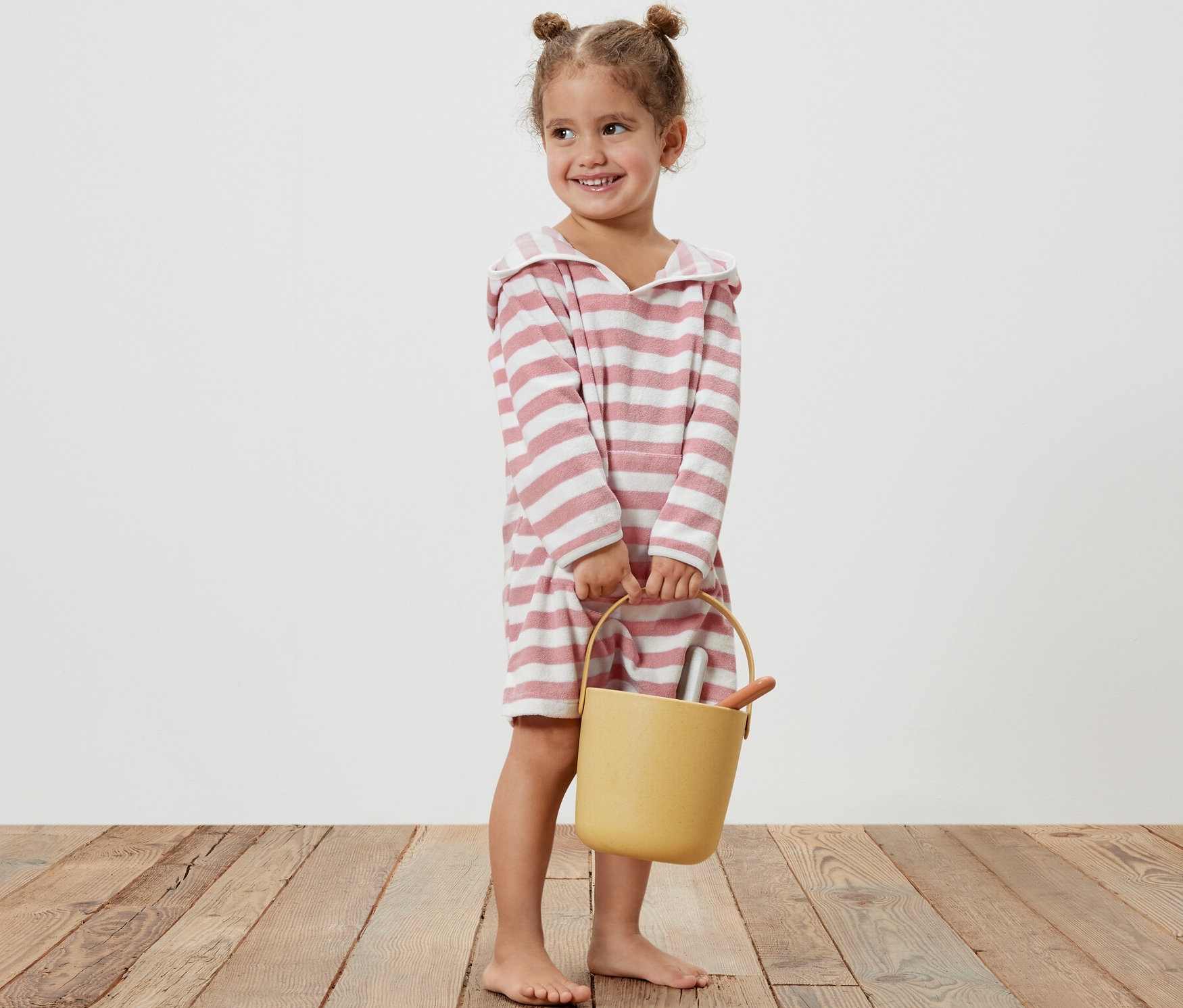 Mosolygó kislány sárga vödröt tart, és rózsaszín csíkos gyermek frottírponcsót visel.