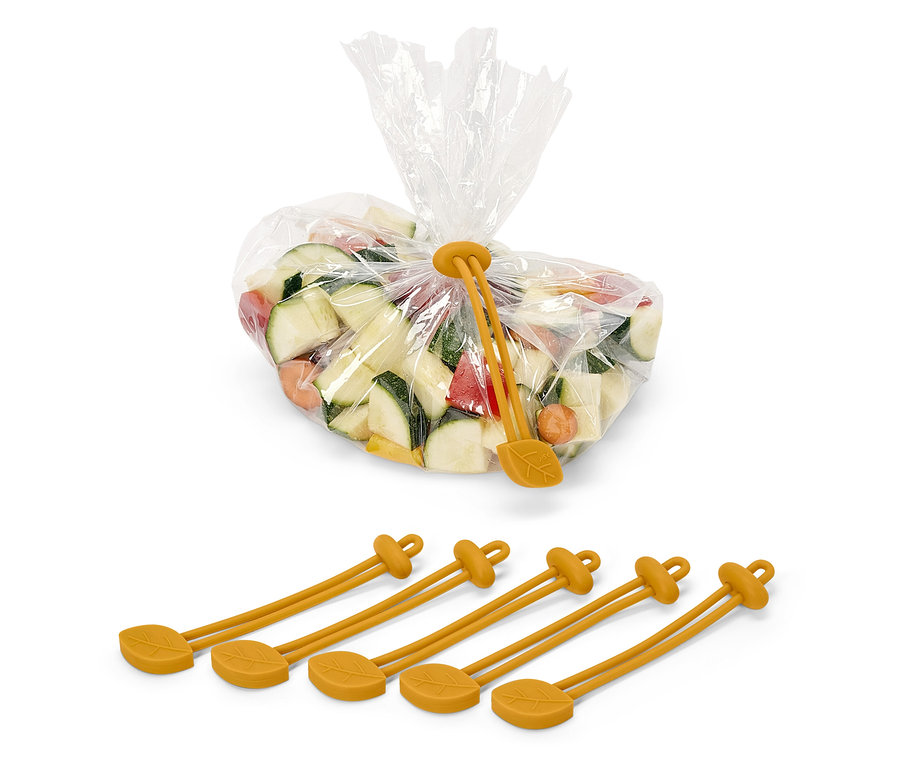 Kockára vágott cukkini, sárgarépa és paprika egy műanyag zacskóban, amelyet egy sárga, levél mintázatú szilikon szorító pánt zár le. A zacskó alatt négy sárga szilikon pánt található.