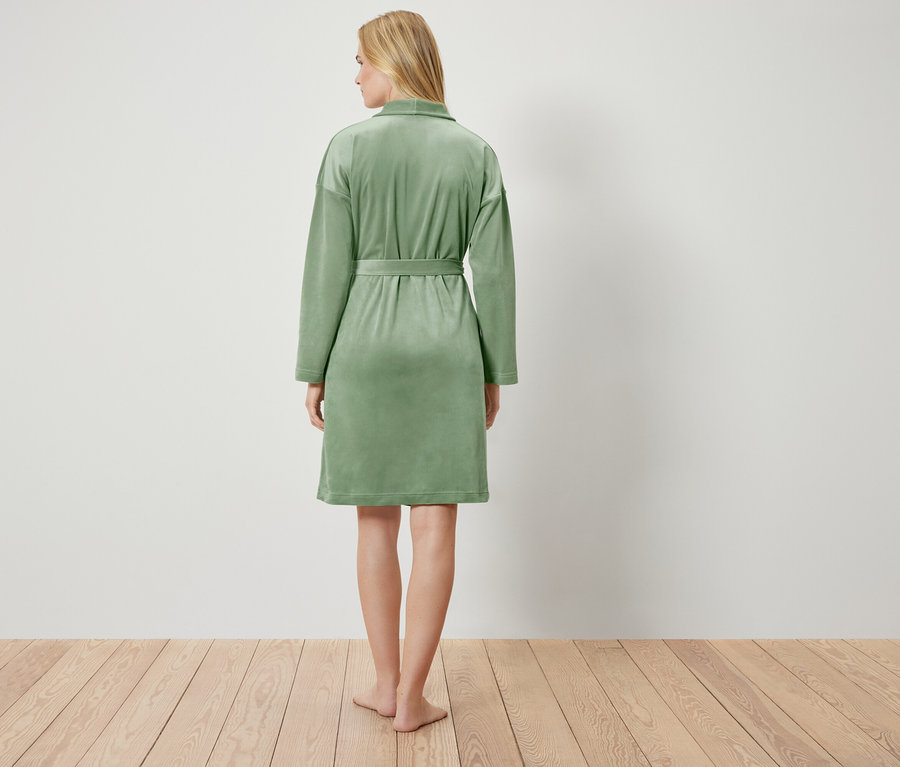 Hátulnézet egy szőke hajú nőről, aki világoszöld köntöst visel, és egy fehér fal előtt áll a fapadlón.