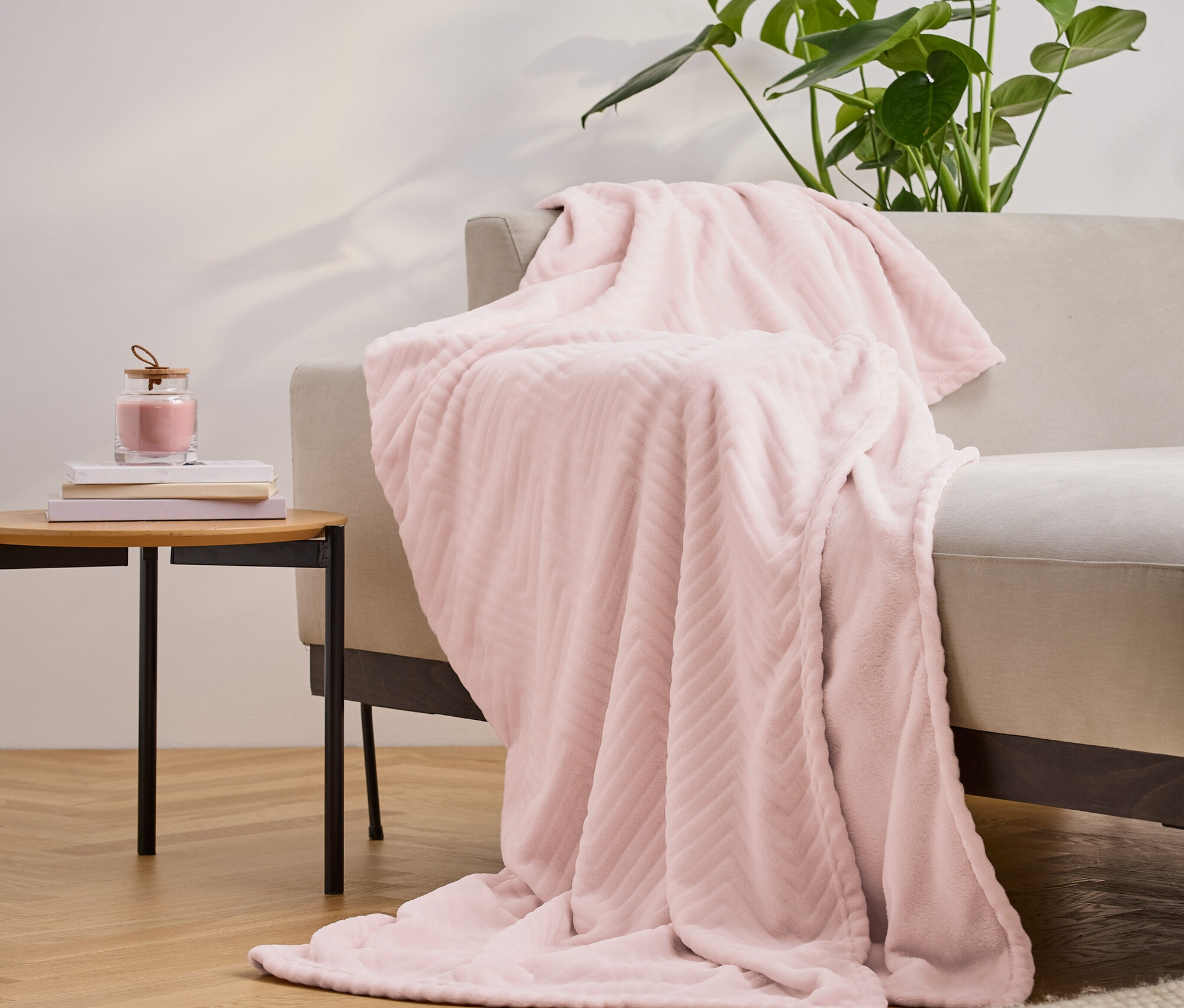 Egy rózsaszín, texturált takaró van átdobva egy bézs kanapén, mellette egy asztal könyvekkel és egy gyertyával, a háttérben pedig egy növény.