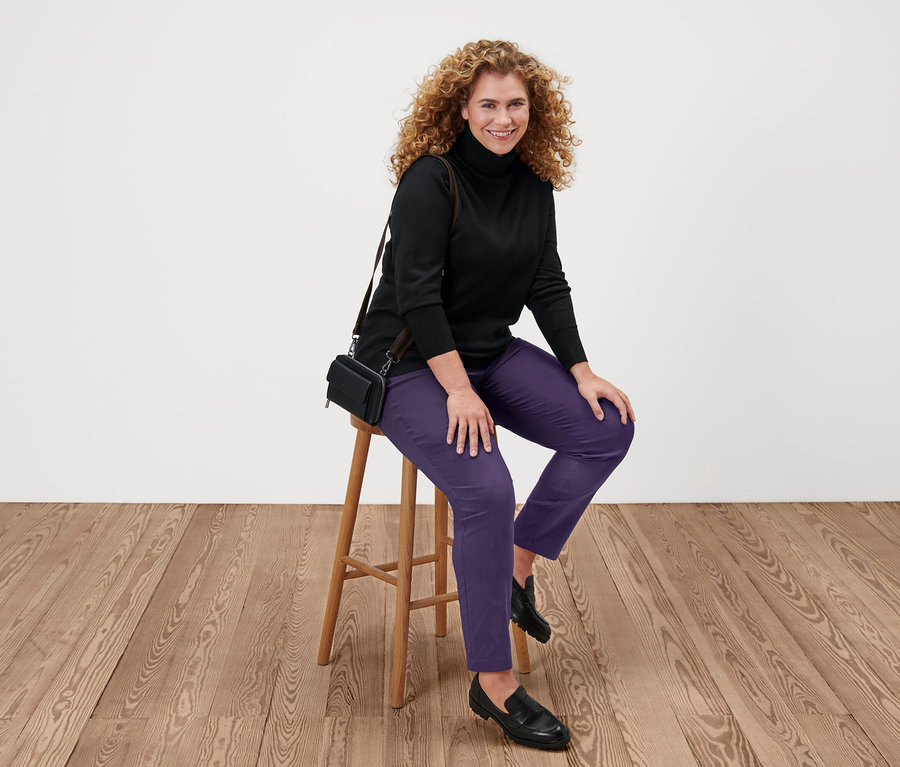 Nő ül egy széken, fekete finom kötésű garbót, lila nadrágot és fekete cipőt visel.