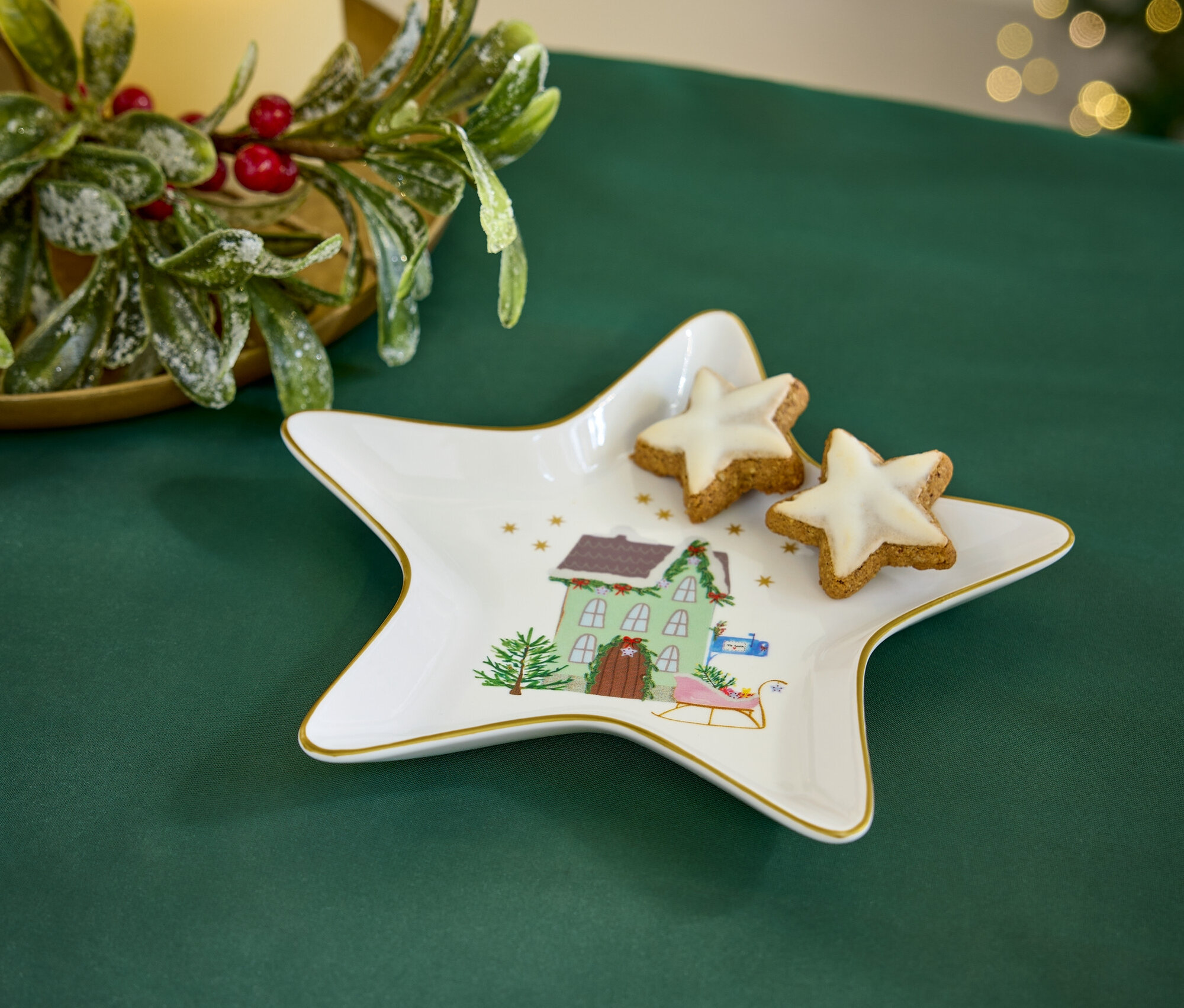 Egy zöld terítőn egy csillag alakú tányér karácsonyi motívummal és két csillag alakú sütemény található. A háttérben egy gyertya és karácsonyi dekoráció látható.