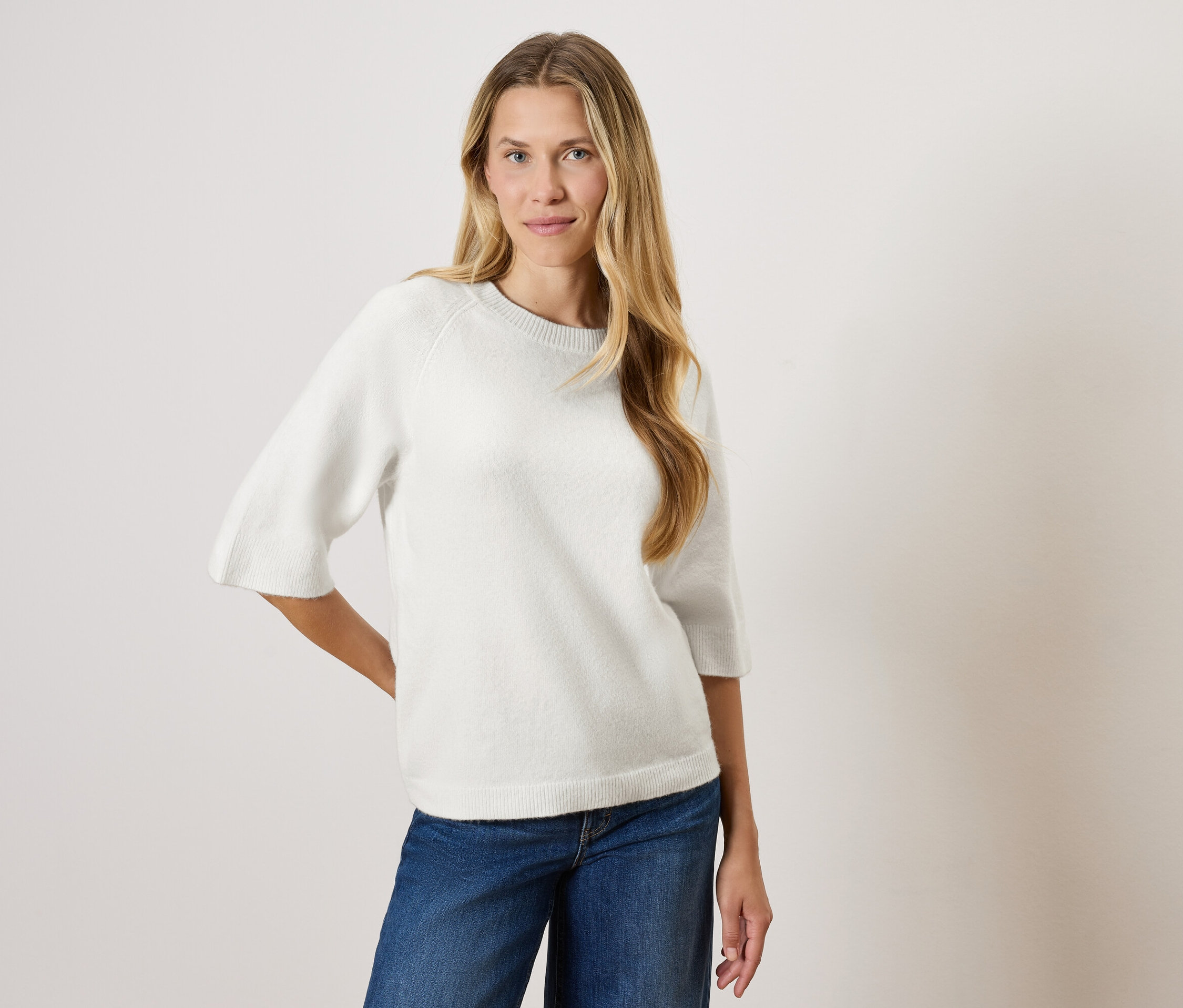 Egy hosszú szőke hajú nő pózol egy fehér, háromnegyedes ujjú pulóverben és kék farmerben.