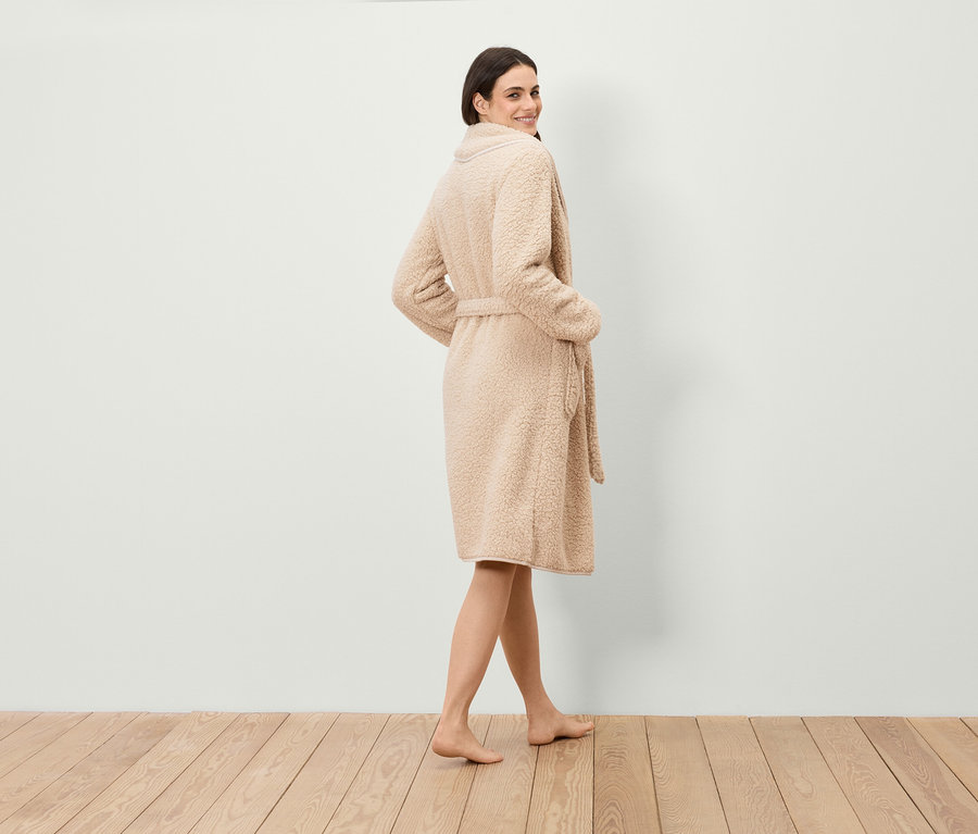 Egy nő a válla fölött hátra néz, műszőrme köntöst visel övvel, és fapadlón áll.