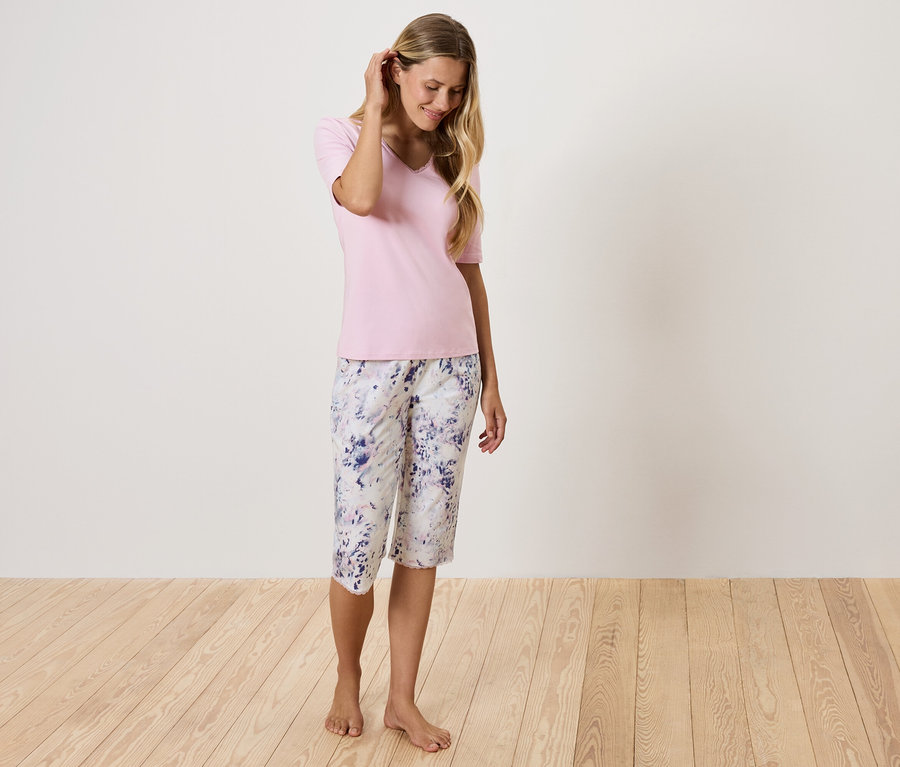 Nő rózsaszín pólóban és virágos nadrágban áll a fapadlón.