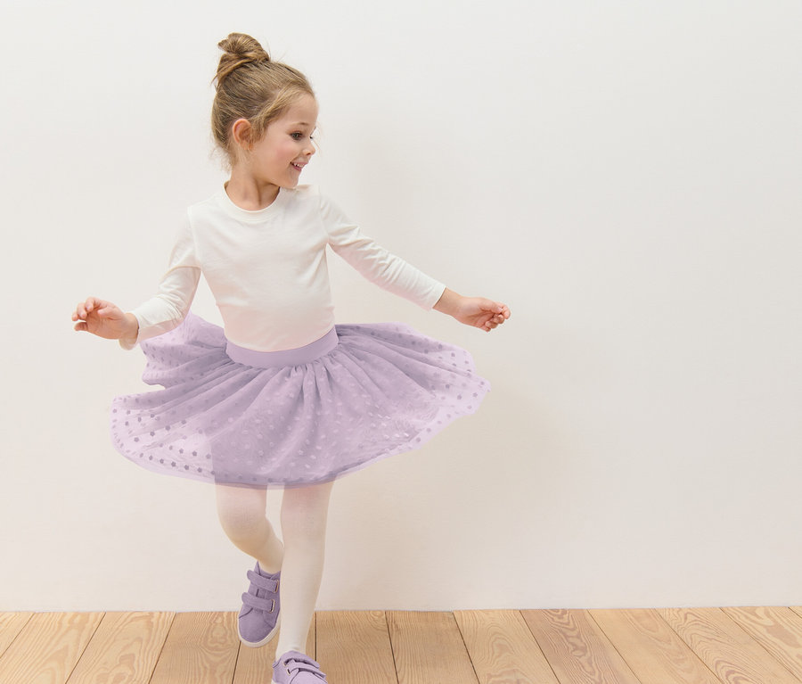 Lány konttyal táncol lila tüllszoknyában, fehér pöttyökkel, hosszú ujjú fehér felsőben, harisnyában és lila cipőben, fehér fal előtt.