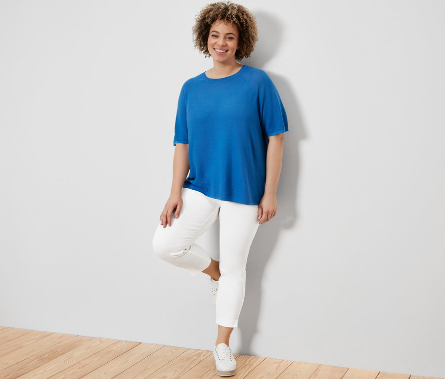 Nő pózol kék finomkötésű felsőben és fehér 7/8-os sztreccsnadrágban.