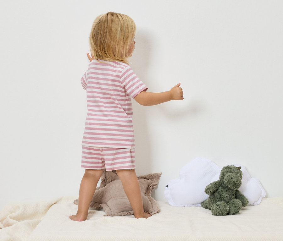 Egy szőke hajú gyermek rózsaszín-fehér csíkos pizsamában áll egy bézs takarón egy zöld dinoszaurusz plüssállat és egy fehér felhőpárna mellett.