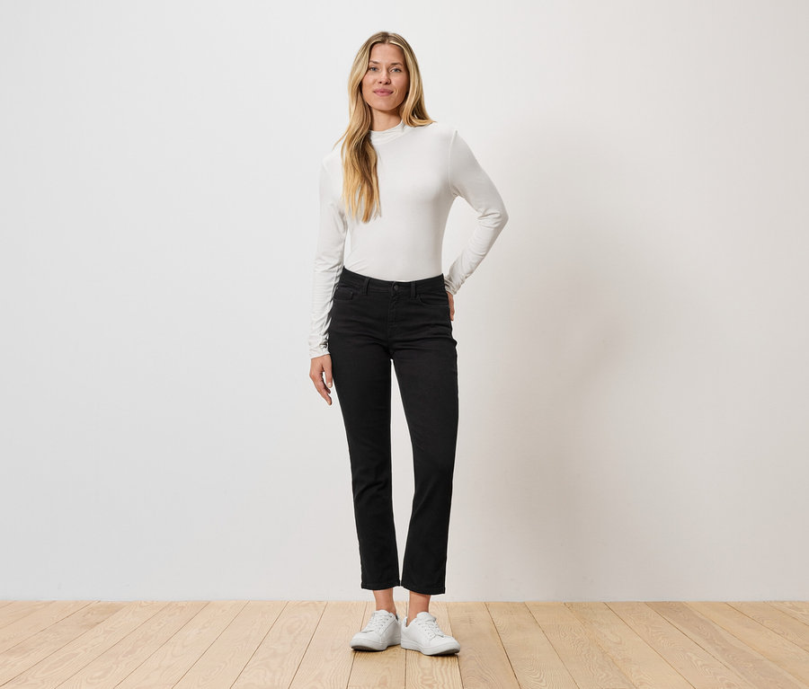 Egy nő áll a fal előtt, hosszú ujjú felsőt, fekete farmert és fehér tornacipőt visel.