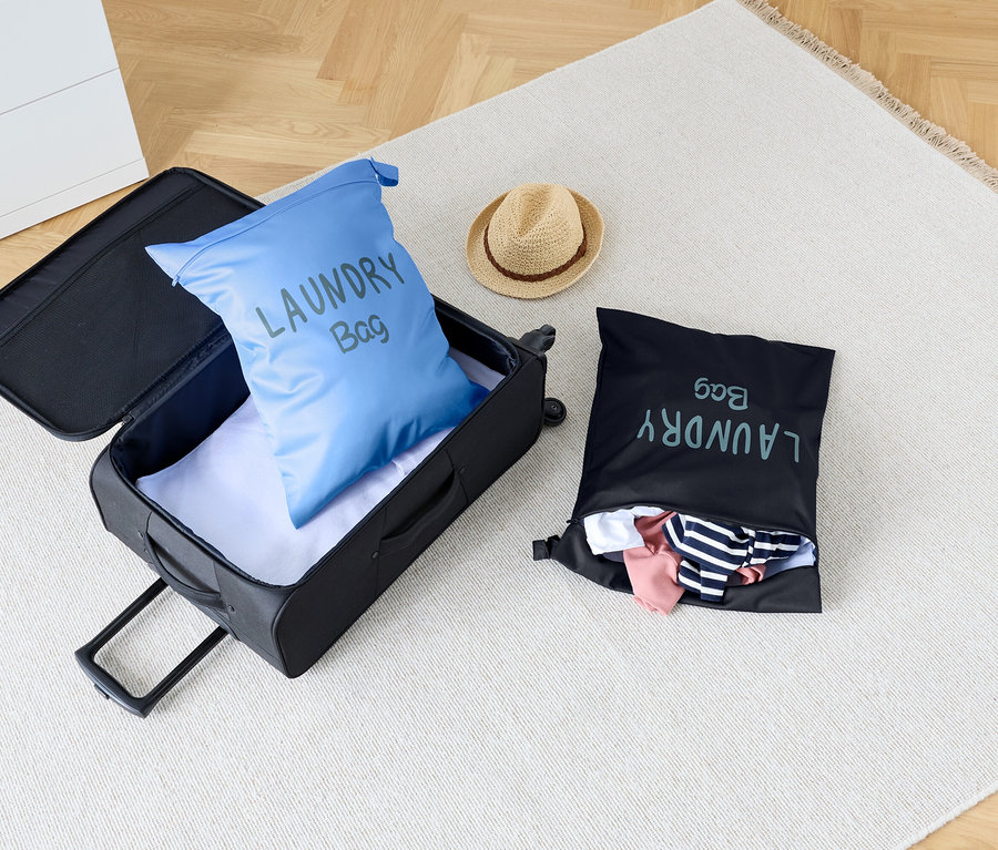 A szőnyegen egy nyitott bőrönd áll, benne egy kék szennyeszsákkal, mellette egy fekete szennyeszsák és egy kalap.